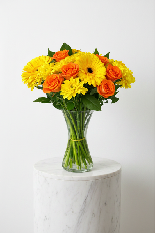 Sunset Delight floral arrangement in a glass vase featuring vibrant orange and yellow flowers with greenery.