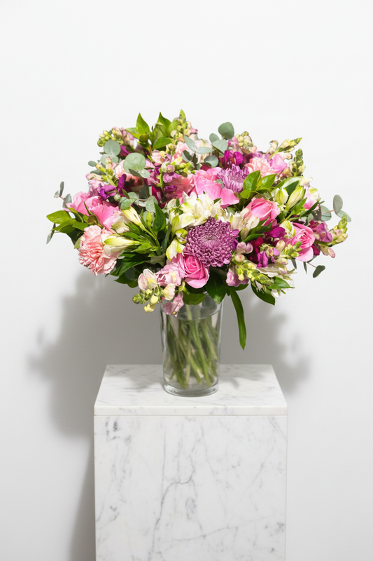Colorful mixed floral bouquet with vibrant flowers arranged in a clear glass vase, displayed on a neutral background.
